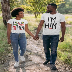 Puede incluir: Una pareja caminando de la mano al aire libre. La mujer lleva una camiseta blanca con el texto "I PRAYED FOR A MAN LIKE HIM" en letras de colores. El hombre lleva una camiseta blanca con el texto "I'M HIM" en letras negras.