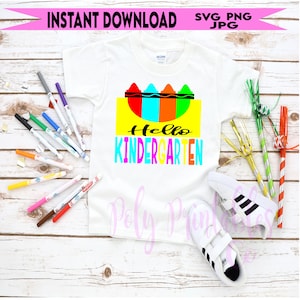 Puede incluir: Una camiseta blanca con el texto "Hello Kindergarten" en letras de colores y un diseño de lápices de colores encima del texto. La camiseta está sobre una superficie de madera blanca con marcadores y artículos de fiesta alrededor.