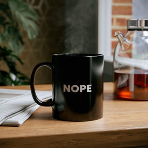 May include: A black ceramic mug with a white handle and the word "NOPE" printed in white on the front. The mug is filled with a hot beverage and steam is rising from the top.
