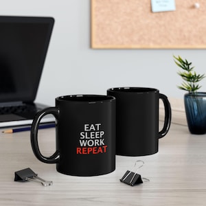 以下が含まれることがあります： 「EAT SLEEP WORK REPEAT」の文字が白と赤でプリントされた、黒いセラミックマグカップが2つ。明るい色の木製の表面に置かれており、背景にはノートパソコンと事務用品があります。