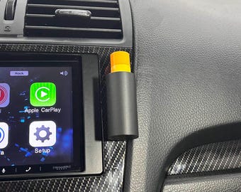 May include: A black and orange car air freshener hanging from the dashboard of a car. The air freshener is attached to a black vent clip. The car's dashboard is black with a carbon fiber pattern.