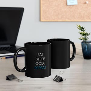Peut inclure: Deux tasses en céramique noire avec du texte blanc. La tasse de gauche a le texte "Eat Sleep Code Repeat" avec le mot "Code" en bleu. La tasse de droite est vide.