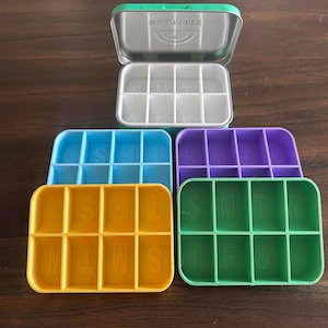 May include: An open, green-lidded metal tin with a divided interior sits above five colorful, plastic pill organizers. The organizers are blue, purple, yellow, and green, each labeled with the days of the week.