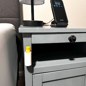 May include: A gray nightstand with a drawer and a door. A small white tube with a yellow top is attached to the side of the nightstand. A black smartphone is on a wireless charging pad on top of the nightstand. The phone screen displays the time 9:44.