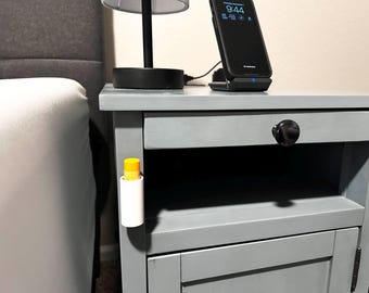 May include: A gray nightstand with a drawer and a door. A small white tube with a yellow top is attached to the side of the nightstand. A black smartphone is on a wireless charging pad on top of the nightstand. The phone screen displays the time 9:44.