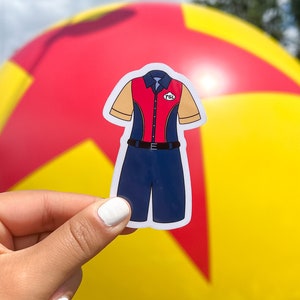 May include: A sticker featuring a cartoon-style jumpsuit with a red and navy blue top, tan sleeves, and navy blue shorts. The sticker has a white border and is held in front of a large yellow, red, and blue ball.