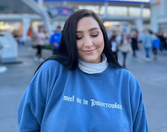 May include: A woman wearing a blue sweatshirt with the text "meet me in Tomorrowland" in white lettering. She is standing in front of a blurred background of a theme park.