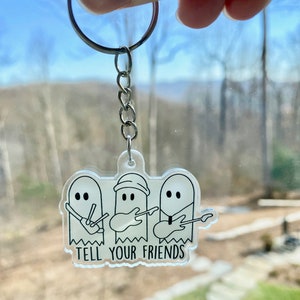 Puede incluir: Un llavero de acrílico transparente con tres fantasmas de dibujos animados tocando instrumentos musicales. Los fantasmas están delineados en negro y tienen rasgos faciales simples. El texto "TELL YOUR FRIENDS" está impreso debajo de los fantasmas.