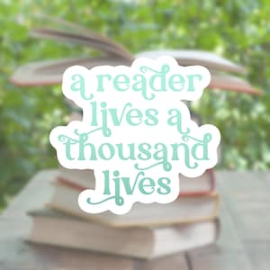 May include: A white sticker with a green quote that reads "a reader lives a thousand lives" on a stack of books with a blurred background of green foliage.