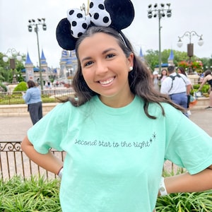 Op de afbeelding: Een vrouw draagt een lichtblauw T-shirt met de tekst "second star to the right*" in zwarte letters. Ze draagt zwarte muizenoren met een witte stippen strik. De vrouw staat voor een kasteel.