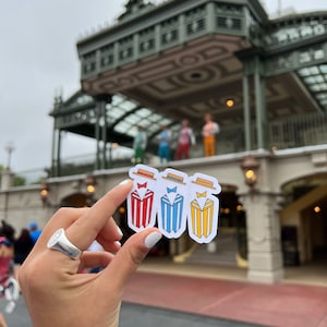 May include: Three cartoon character stickers with red, blue, and yellow striped suits and hats. The stickers are held in front of a building with a green roof.