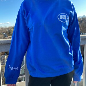 May include: Royal blue sweatshirt with a white logo on the chest and the word "Scarer" embroidered on the sleeve. The sweatshirt has a crew neck. The person is wearing black leggings. The background is a bright, sunny day.