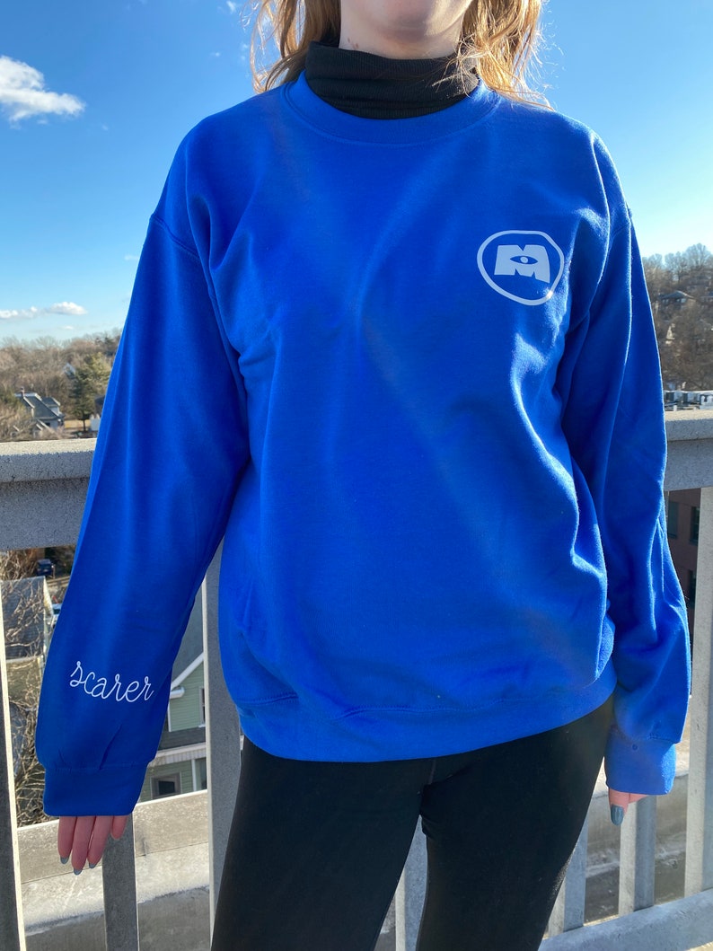 May include: Royal blue sweatshirt with a white logo on the chest and the word "Scarer" embroidered on the sleeve. The sweatshirt has a crew neck. The person is wearing black leggings. The background is a bright, sunny day.