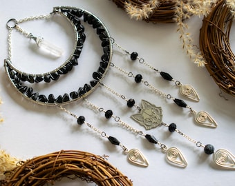 May include: A crescent moon-shaped wall hanging with black stones and silver wire detailing. The design includes dangling chains with black beads, silver teardrop shapes, and a silver cat skull pendant. A clear crystal hangs from the top.