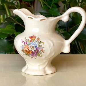 Puede incluir: Una jarra de crema de cerámica blanca con un diseño floral en tonos de rosa, naranja, amarillo, azul y verde. La jarra tiene un borde festoneado y un asa.