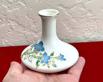 May include: A small white ceramic vase with a gold rim and blue floral design. The vase has a rounded base and a narrow neck.
