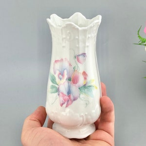 Può includere: Un vaso in ceramica bianca con bordo smerlato e base svasata. Il vaso è decorato con un motivo floreale nei toni del rosa, viola e blu, con foglie verdi. Il vaso è alto circa 15 cm.