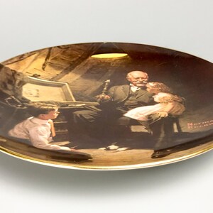 May include: A ceramic plate with a gold rim features a painting of a man with two children. The man is sitting in a chair and the children are standing next to him. The painting is in a dark brown color scheme and the plate has a white background. The artist's signature, "Norman Rockwell", is visible in the lower right corner of the plate.