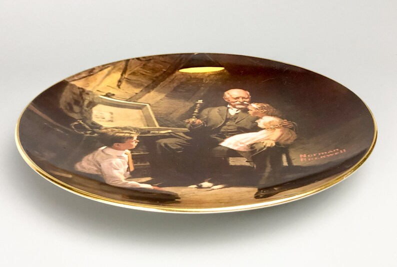 May include: A ceramic plate with a gold rim features a painting of a man with two children. The man is sitting in a chair and the children are standing next to him. The painting is in a dark brown color scheme and the plate has a white background. The artist's signature, "Norman Rockwell", is visible in the lower right corner of the plate.