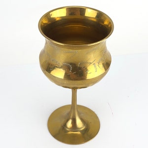 May include: A vintage brass goblet with a wide bowl and a slender stem. The goblet features etched floral designs around the bowl. The base is a wide, circular foot. The goblet has a polished, golden finish.