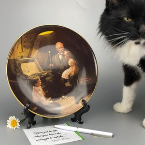 May include: A decorative ceramic plate featuring a painting of a man and two children looking at a treasure chest. The plate has a gold rim and is on a black stand. The quote "You get in life what you have the courage to ask for." is written on a notecard next to the plate.
