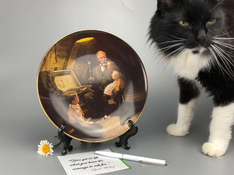 May include: A decorative ceramic plate featuring a painting of a man and two children looking at a treasure chest. The plate has a gold rim and is on a black stand. The quote "You get in life what you have the courage to ask for." is written on a notecard next to the plate.
