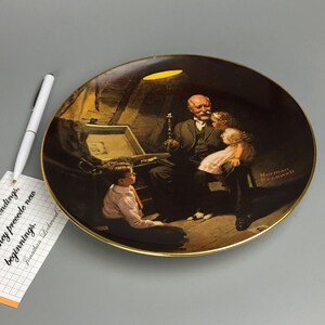 May include: A porcelain plate with a gold rim features a painting of a man with two children. The man is holding a candle and looking at the children. The painting is set in a dimly lit room with a wooden chest in the background. The plate has the artist's signature "Norman Rockwell" in the bottom right corner.
