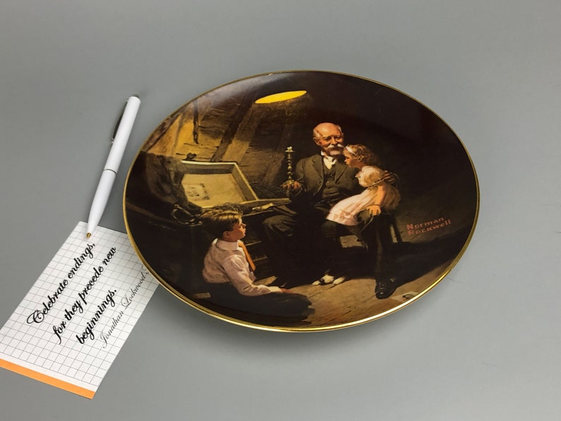 May include: A porcelain plate with a gold rim features a painting of a man with two children. The man is holding a candle and looking at the children. The painting is set in a dimly lit room with a wooden chest in the background. The plate has the artist's signature "Norman Rockwell" in the bottom right corner.