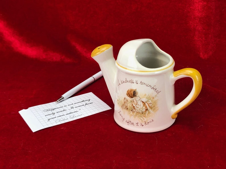 Pode incluir: Um regador de cer&acirc;mica branca com uma pega e um bico amarelos. Tem um desenho floral e a cita&ccedil;&atilde;o "A kindness is remembered long after it is done".
