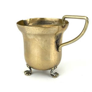 Op de afbeelding: Een kleine, antieke messing beker met een handvat en drie decoratieve voetjes. De beker heeft een licht uitlopende rand en een aangetaste afwerking. Het rechthoekige handvat is aan de zijkant bevestigd. De sierlijke voetjes dragen bij aan de vintage esthetiek.