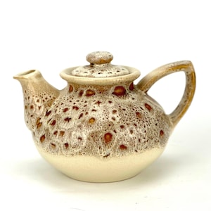 Può includere: Una teiera in ceramica con smalto maculato marrone e crema. La teiera ha un corpo arrotondato, un manico curvo e un coperchio con un piccolo pomello. Il beccuccio è corto e angolato. La teiera è di colore neutro.