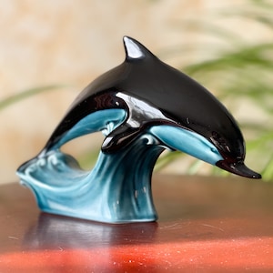 Può includere: Una statuetta di delfino in ceramica nera e blu che salta fuori da un'onda. Il delfino è lucido e ha un design realistico.