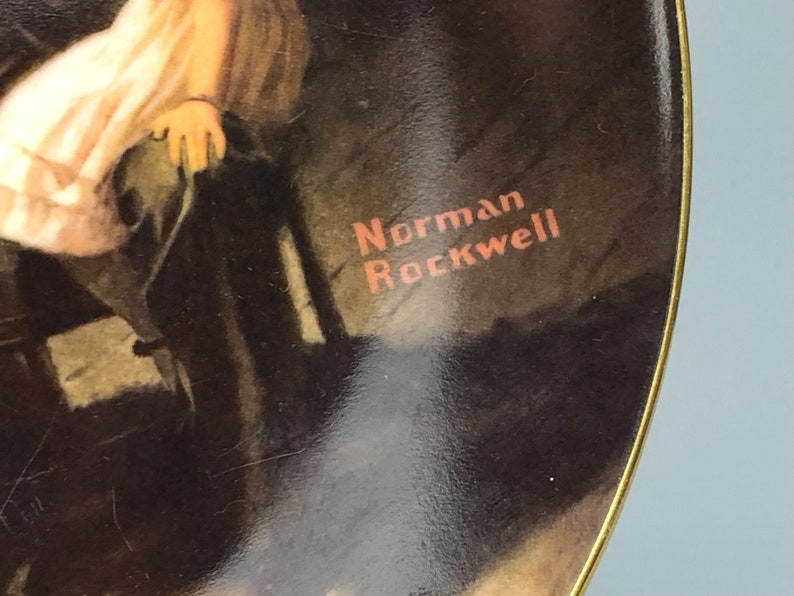 May include: A round ceramic plate with a gold rim features a painting of a young girl in a white dress sitting on a chair. The plate has the artist's signature, "Norman Rockwell", printed in red on the bottom right corner.