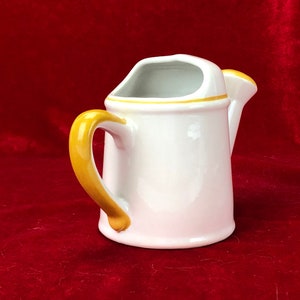 Pode incluir: Um regador de cer&acirc;mica branca com uma al&ccedil;a amarela e uma borda amarela. O regador est&aacute; sobre uma superf&iacute;cie de veludo vermelho.