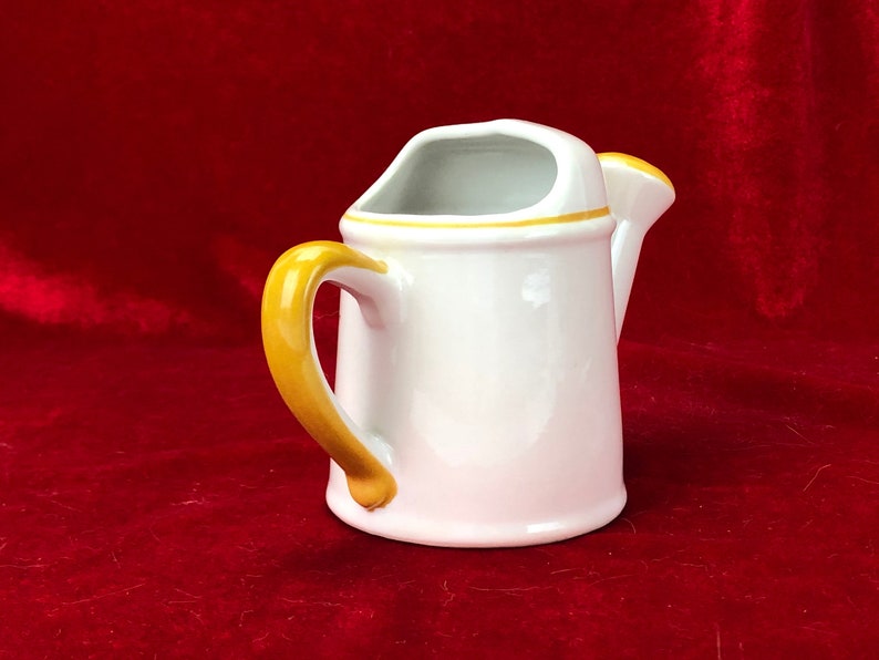 Pode incluir: Um regador de cer&acirc;mica branca com uma al&ccedil;a amarela e uma borda amarela. O regador est&aacute; sobre uma superf&iacute;cie de veludo vermelho.