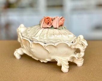Puede incluir: Joya de cerámica de color crema con tapa, con un diseño floral y tres pequeñas rosas color melocotón en la parte superior. La caja tiene detalles ornamentados y está sobre pequeños pies.