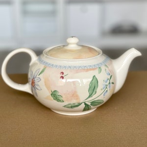 Op de afbeelding: Een crèmekleurige theepot van keramiek met een handgeschilderd bloemmotief van groene bladeren, blauwe bloemen en kleine rode bessen. De theepot heeft een bijpassend deksel met knop, een gebogen tuit en een blauwe rand met een kruispatroon.