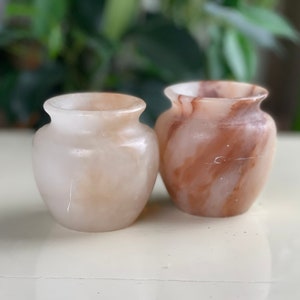 Puede incluir: Dos pequeños jarrones decorativos. El jarrón de la izquierda es de color crema pálido, mientras que el de la derecha tiene una mezcla de tonos marrones y crema. Ambos jarrones tienen un cuerpo redondeado y un cuello estrecho.