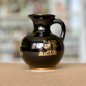 May include: A small, black ceramic jug with a handle and a gold rim. The jug features a gold illustration of figures in a classical style. The jug is likely used for serving liquids and has a glossy finish.