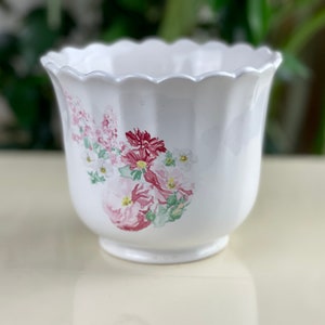 May include: White ceramic planter with a scalloped rim and a floral design featuring pink, red, and green flowers.