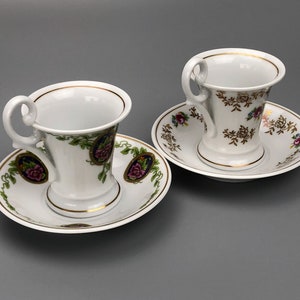 以下が含まれることがあります： 金色の縁取りと花柄の白い磁器製のティーカップとソーサー2つ。カップは広がった形をしていて、持ち手があります。ソーサーは花柄で、金色のアクセントが施されています。