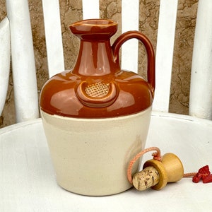 Puede incluir: Una botella de cerámica marrón y blanca con un tapón de corcho y una cuerda. La botella tiene un asa y un diseño decorativo en la parte delantera.