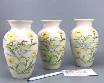 Könnte beinhalten: Drei cremefarbene Keramikvasen mit handgemaltem Design aus gelben Blumen, grünem Laub und kleinen Häusern auf einem Hügel. Die Vasen haben einen leicht ausgestellten Rand und eine glatte, glänzende Oberfläche.