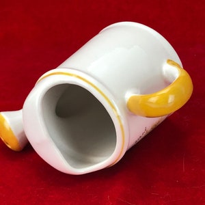 Pode incluir: Regador de cer&acirc;mica branca com al&ccedil;a amarela. O regador &eacute; decorado com uma borda amarela e um desenho floral.