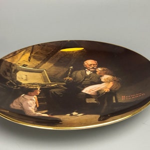 May include: A vintage ceramic plate featuring a Norman Rockwell painting of an older man reading to two children. The painting is set in a dimly lit room with a single light source illuminating the scene. The plate has a gold rim.