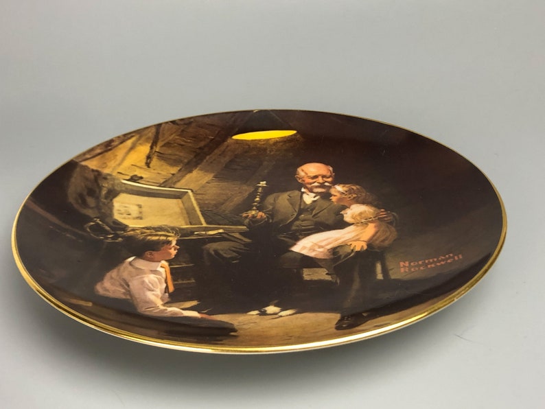 May include: A vintage ceramic plate featuring a Norman Rockwell painting of an older man reading to two children. The painting is set in a dimly lit room with a single light source illuminating the scene. The plate has a gold rim.