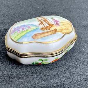 Pode incluir: Uma caixa de porcelana branca com guarnição dourada e uma cena pintada de um veleiro em uma massa de água. A caixa tem forma oval e uma tampa com dobradiças.