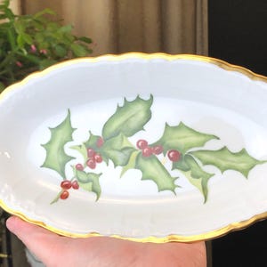 Peut inclure: Plat ovale blanc avec un bord doré, décoré d'un motif de houx et de baies. Les feuilles de houx sont vertes et les baies sont rouges. Une pièce festive pour servir des collations ou de petits plats.