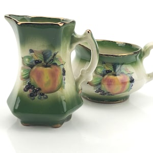 May include: A vintage ceramic creamer and sugar bowl set. The pieces are green with a gold trim and feature a painted design of apples, grapes, and leaves. The creamer has a handle and a spout, while the sugar bowl has a handle.
