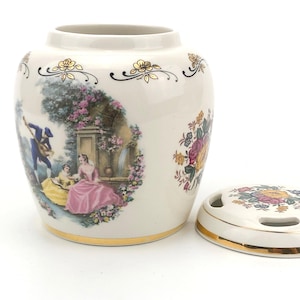 Puede incluir: Un jarrón de cerámica crema con tapa, con una escena romántica y motivos florales. El jarrón tiene un borde dorado y diseños florales. La tapa tiene un borde dorado y un diseño floral.